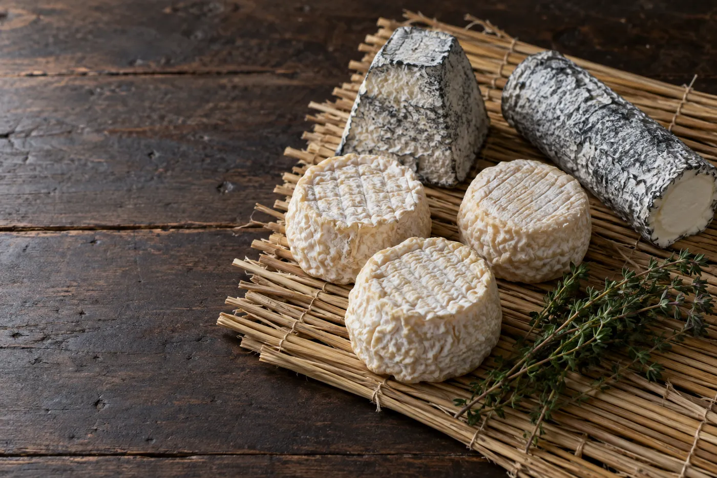 Illustration: kleine Ziegenkäse, eine Pyramide und eine Rolle in Asche auf einer Strohmatte, daneben frischer Thymian